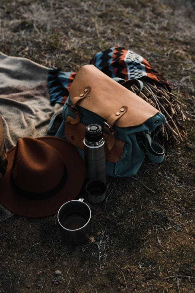 A cozy outdoor picnic layout featuring a backpack, hat, thermos, and blanket on grassy ground.