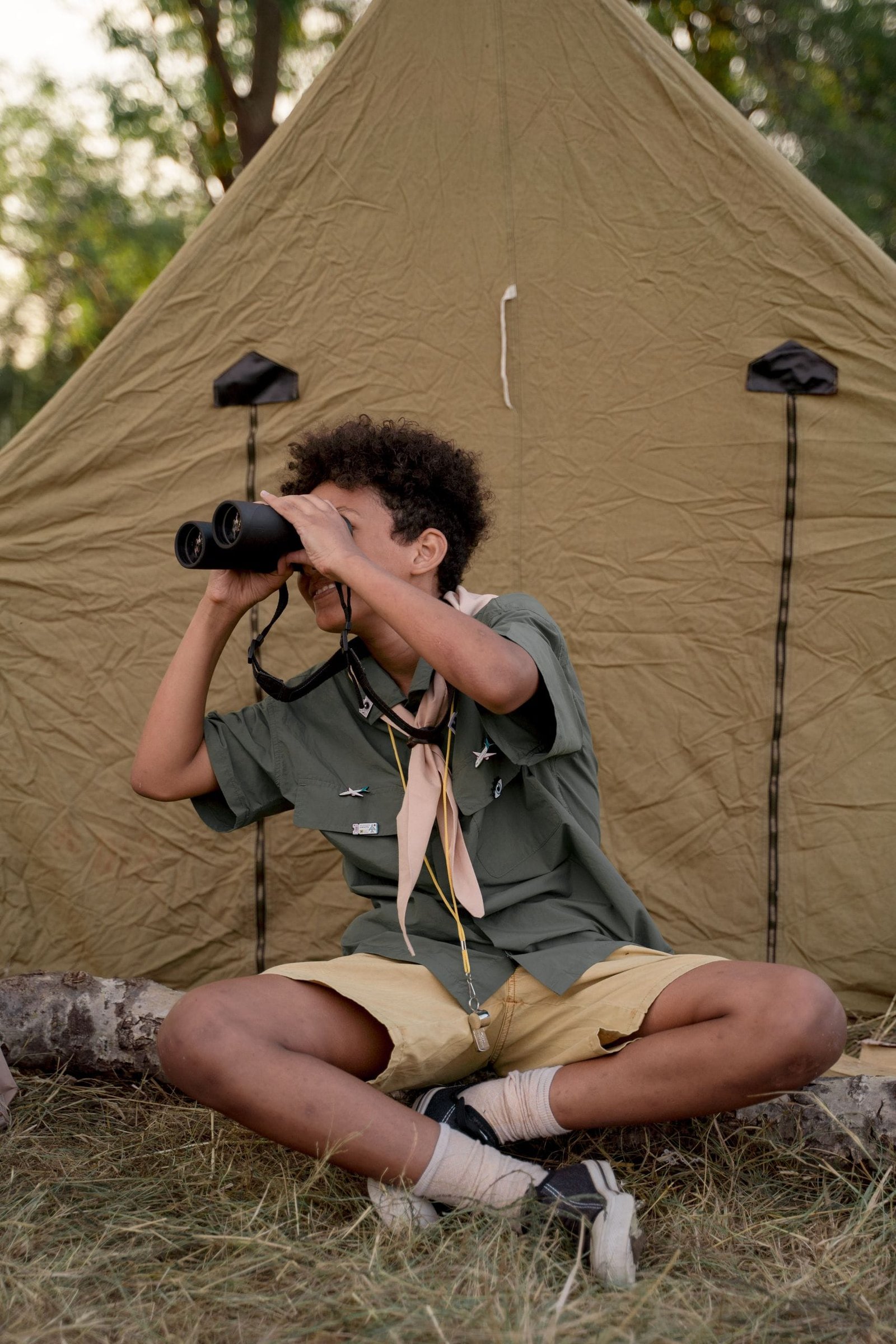 A young boy sits in front of a tent, using binoculars, enjoying an outdoor camping experience.