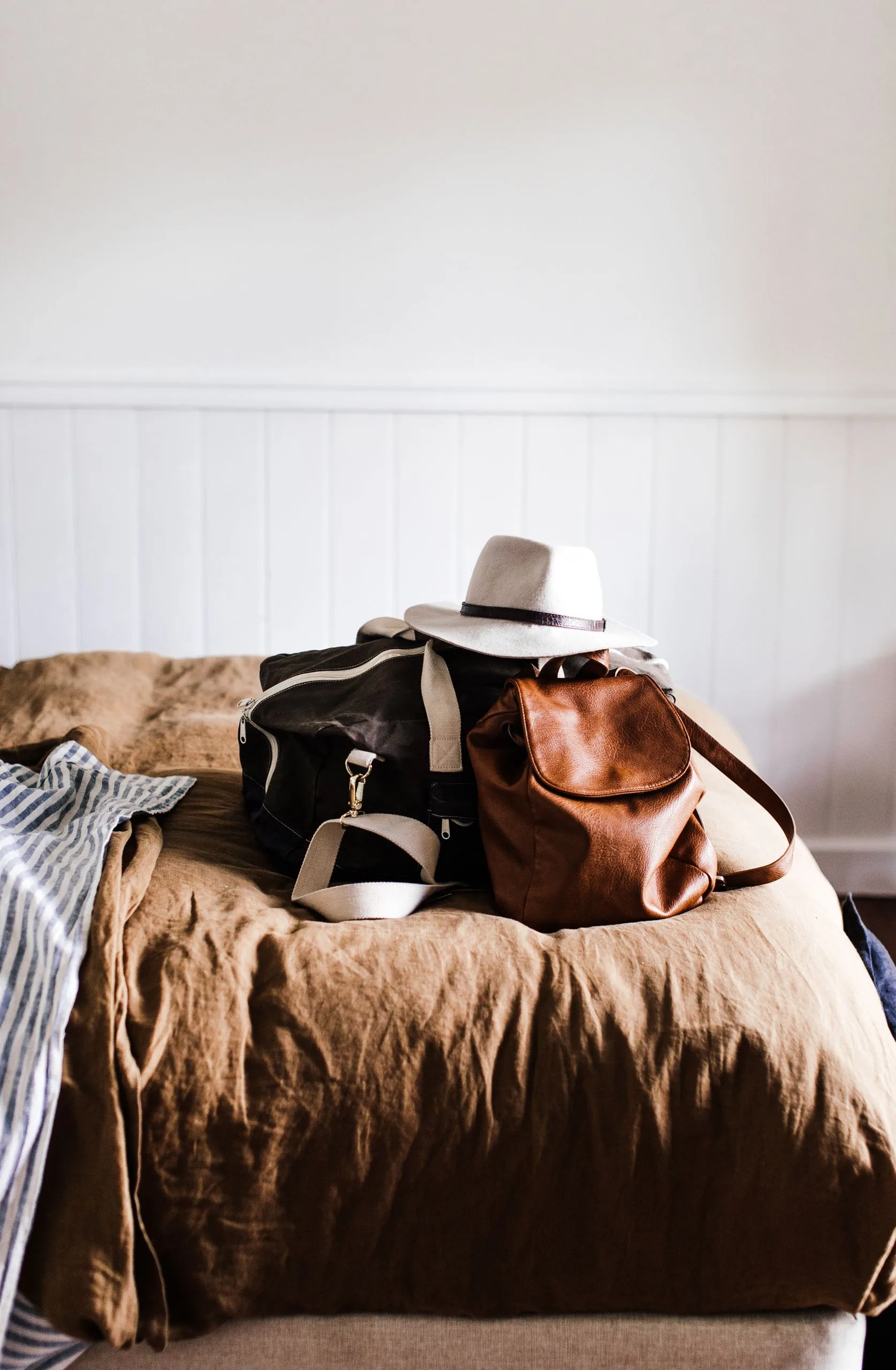 Hat on leather rucksack and travel bag on crumpled bed cover against white wall at home